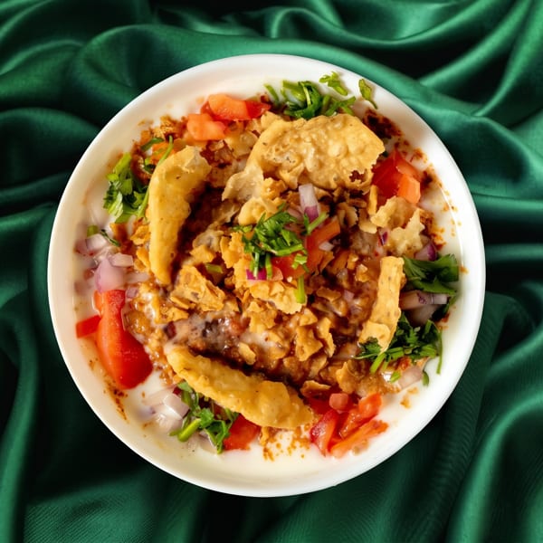 A white bowl of Dahi Bhalla Chaat with yogurt, spices, and crispy toppings on green silk.